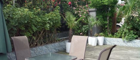 Um pátio externo com mesa e cadeiras, cercado por vegetação exuberante e plantas floridas.