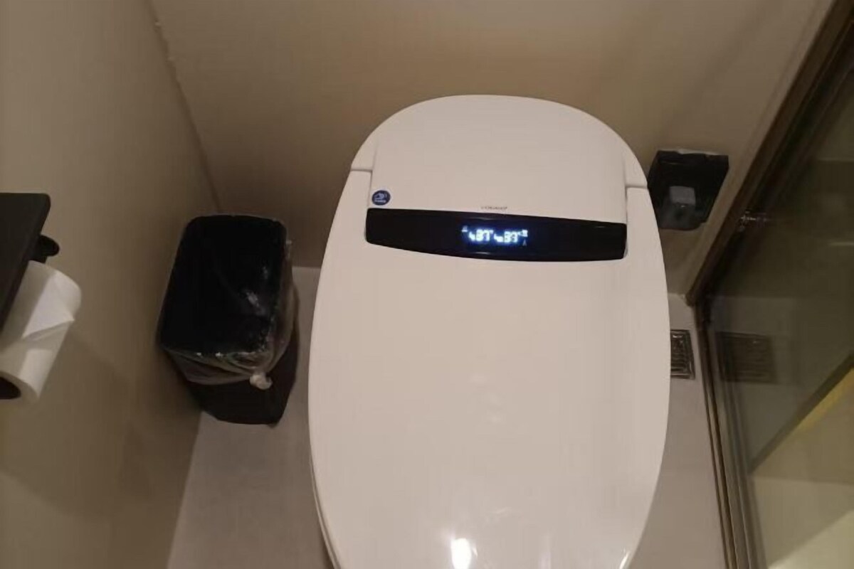A white toilet with a digital display, a trash bin, and a paper towel dispenser.