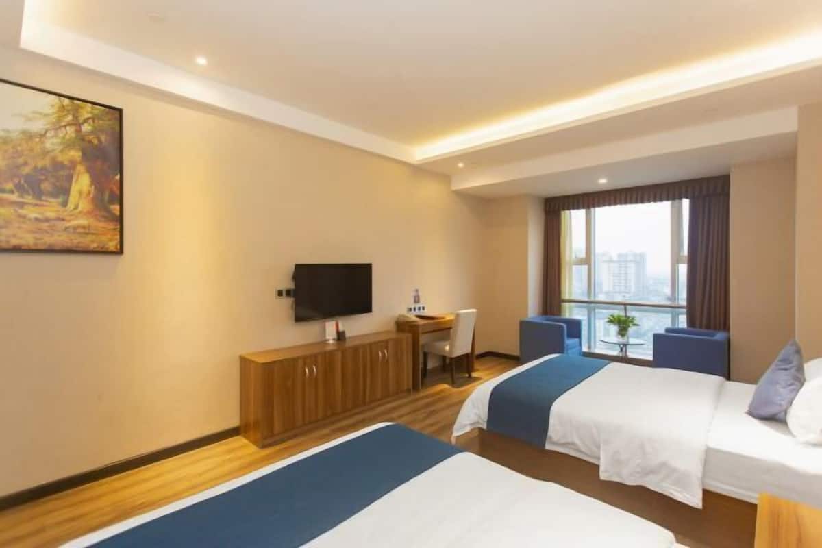 A hotel room with two beds, a desk, a TV, and a large window with a city view.