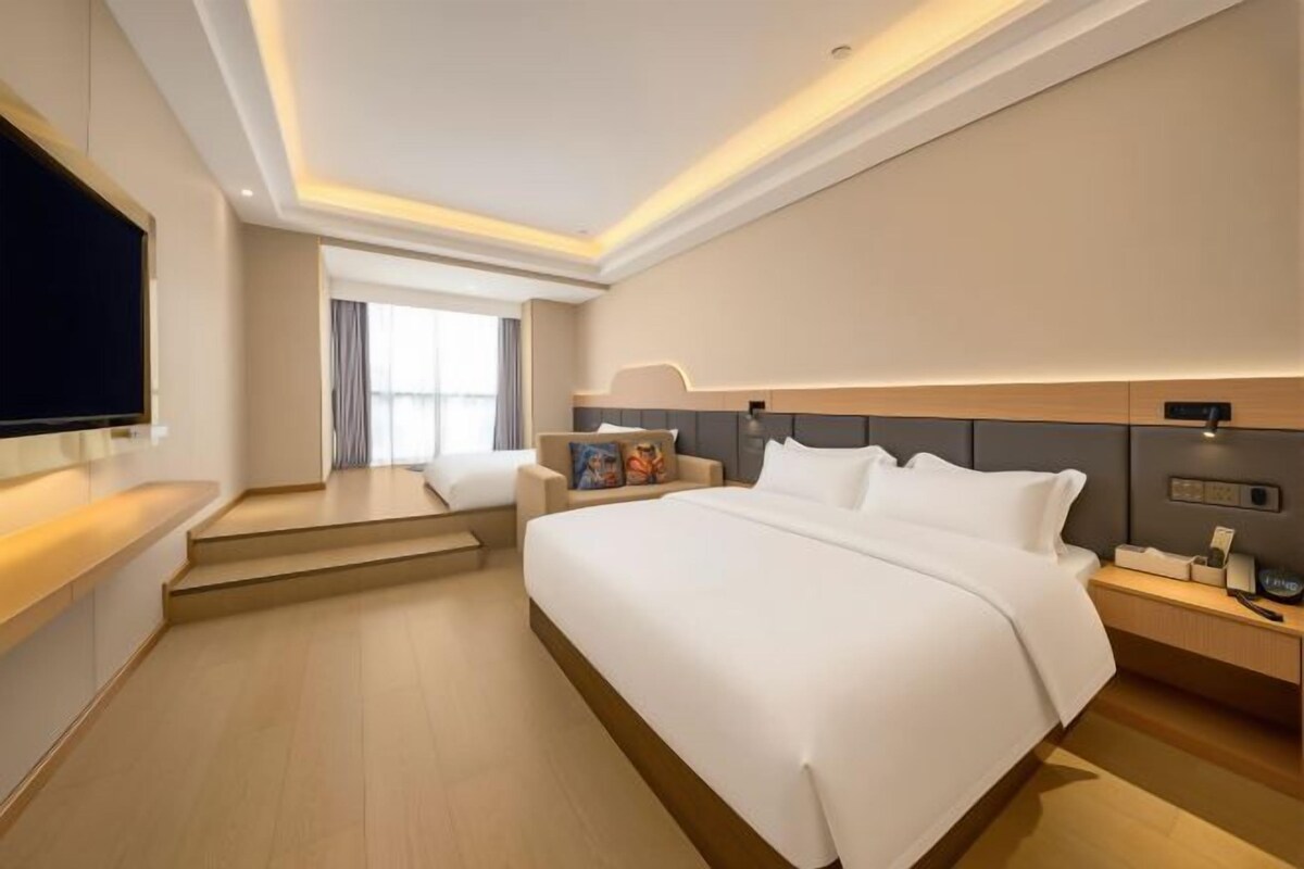 A modern hotel room with a large bed, a flat-screen TV, a bedside table, and a window with curtains.