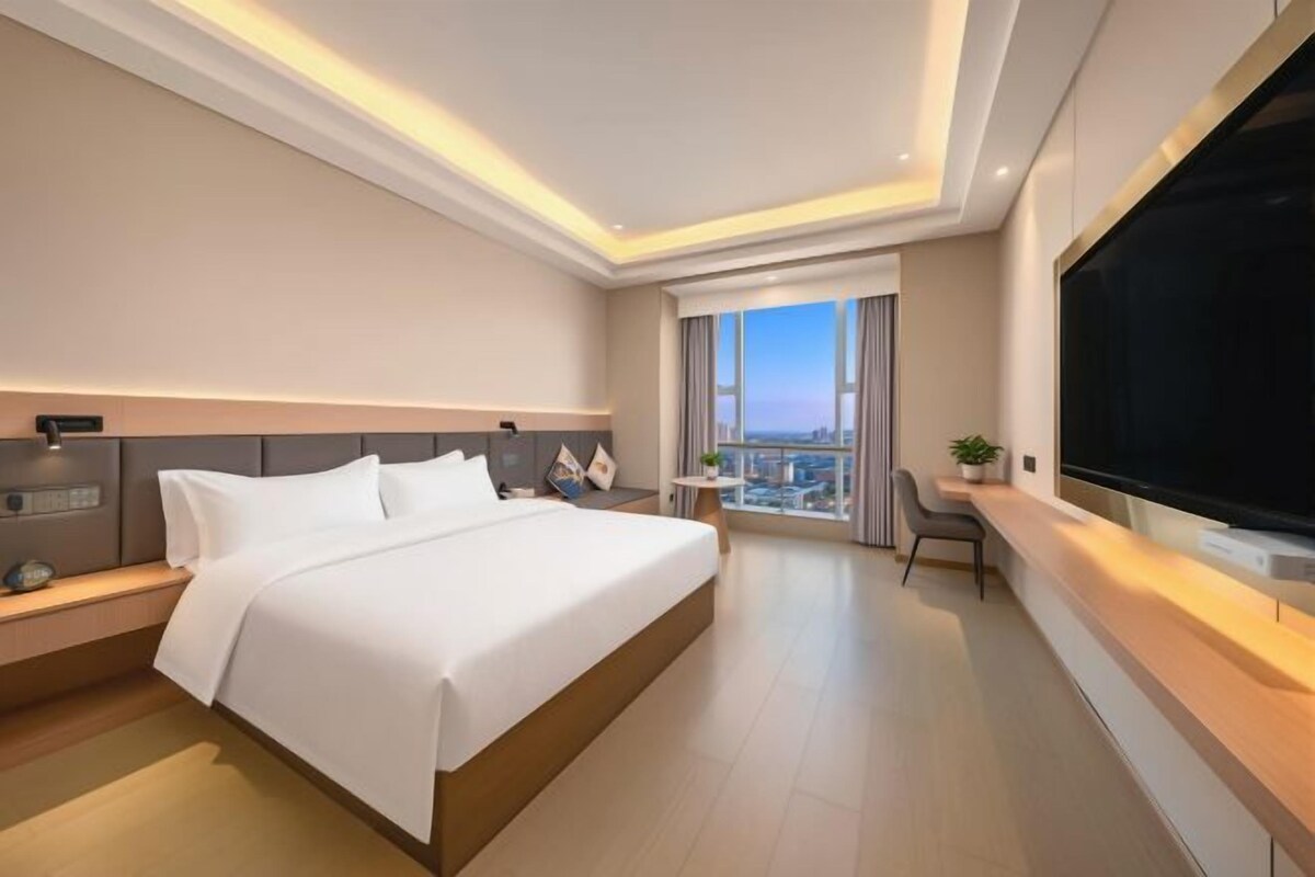 A modern hotel room with a large bed, bedside tables, a flat-screen TV, a city view, and a balcony.