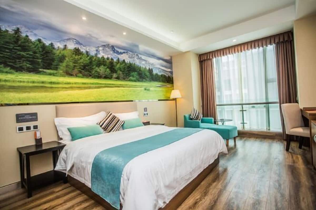 A hotel room with a large bed, a desk, a chair, a small table, a lamp, and a view of a mountain landscape through the window.