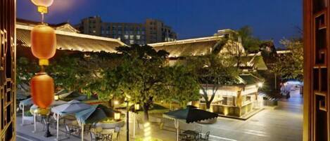 An outdoor dining area with tables and umbrellas, surrounded by trees and buildings in the background.