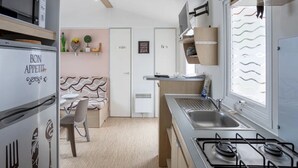 A compact kitchen with a refrigerator, stove, and microwave. There is a dining area with a chair and a small table. The kitchen has a window with blinds and a door labeled "Bain".