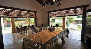 An indoor dining area with a long table covered in a floral tablecloth, surrounded by chairs. There are two large windows with a view of a garden outside.