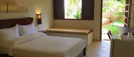A hotel room with a bed, a bench, a wall-mounted light, and a window with a view of greenery.
