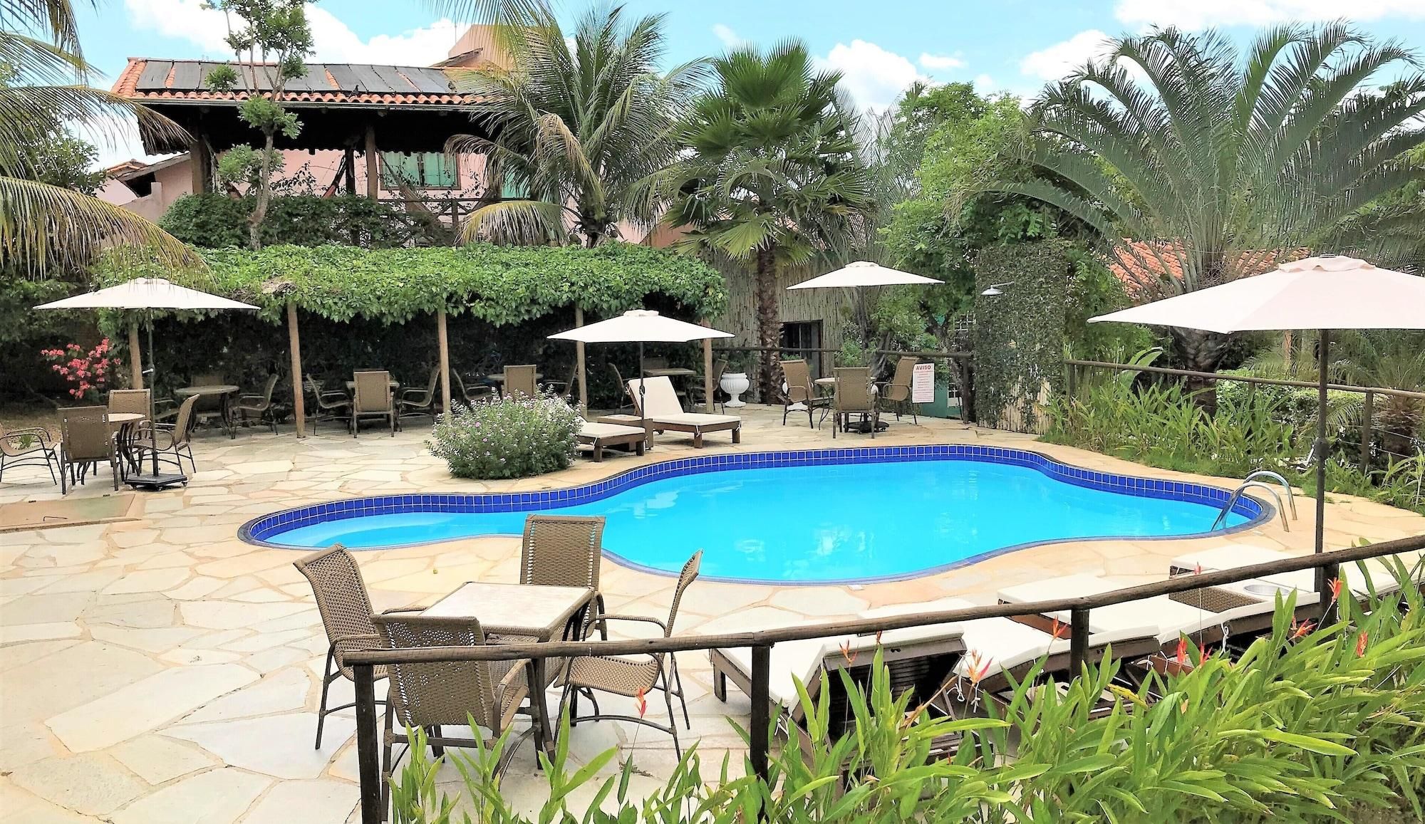 A swimming pool with lounge chairs and umbrellas, surrounded by greenery and a building with a tiled roof.