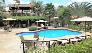 A swimming pool with lounge chairs and umbrellas, surrounded by greenery and a building with a tiled roof.