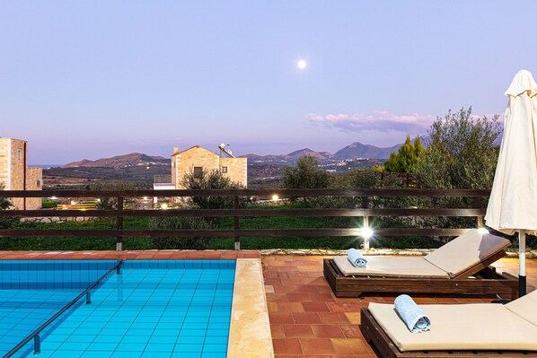 A rooftop pool with clear blue tiles, wooden lounge chairs with towels, and a view of distant mountains.