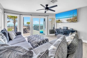 Un salon avec un canapé gris, un ventilateur de plafond et une grande fenêtre donnant sur une plage.