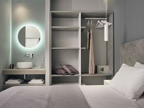 A modern bedroom with a large bed, a built-in wardrobe with hanging space, a bedside table with a lamp, and a round mirror above the sink. - Nikos & Maria Studios II (Zakynthos)