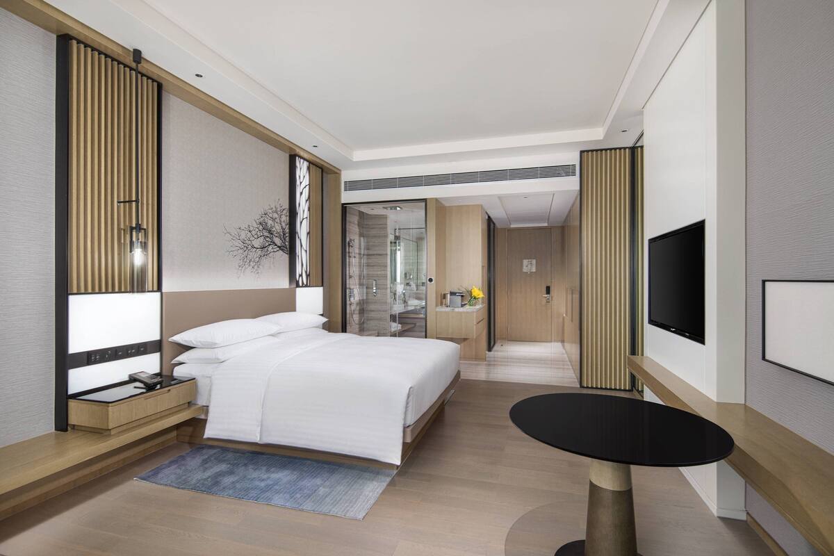 A modern hotel room with a large bed, a desk, a flat-screen TV, and a small round table.