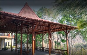 Un pavillon en bois, surmonté d’un toit en tuiles, entouré d’arbres et avec une chaîne de montagnes en arrière-plan.