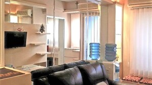 A living room with a black leather sofa, a flat-screen TV mounted on the wall, a water dispenser, and a large window with blinds. - Apartement Puncak Permai Tower A by Nur Faizah (Surabaya)