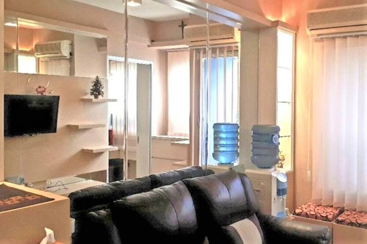 A living room with a black leather sofa, a flat-screen TV mounted on the wall, a water dispenser, and a large window with blinds.