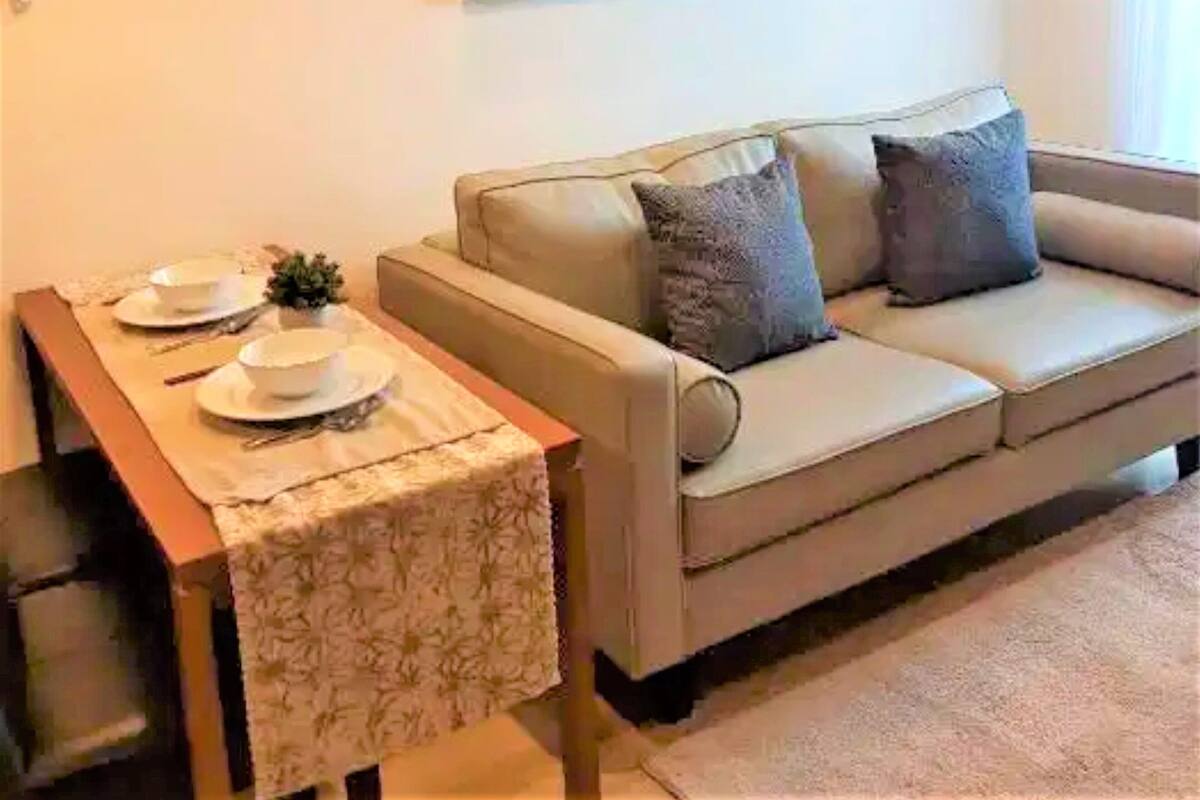 Ein Wohnzimmer mit einem beigefarbenen Sofa, zwei Kissen, einem kleinen Tisch mit Tischdecke und einem Wandbild.