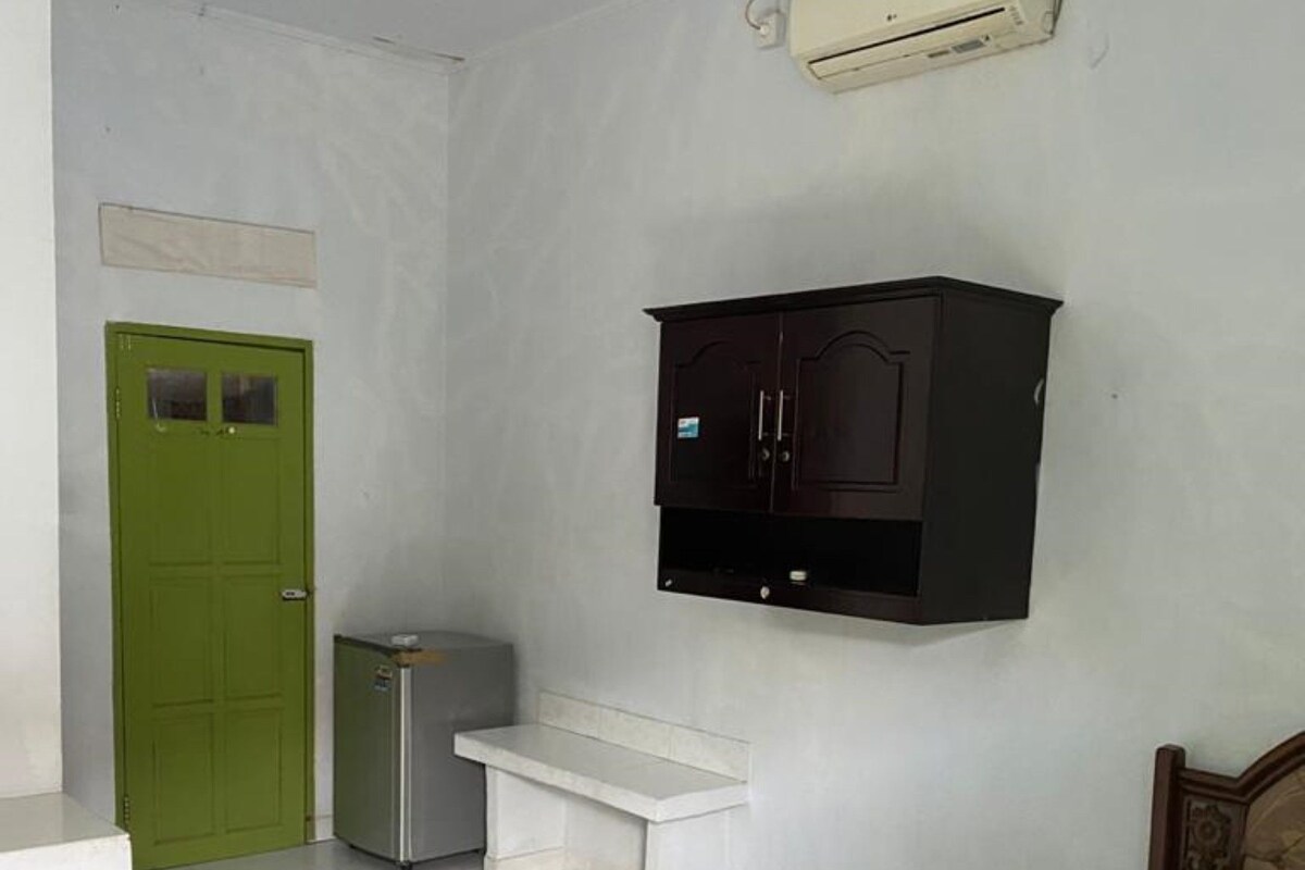 A bedroom with a bed, a green door, a white bench, a small refrigerator, and a wall-mounted cabinet.