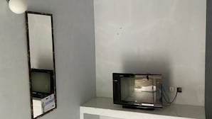 A room with a white tiled floor, a white bench, a black television on a stand, and a wall-mounted mirror with a light fixture. - Wisma Adhyaksa 2 (Makassar)