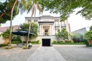 A building with a central entrance, surrounded by a paved courtyard, palm trees, and a covered seating area.