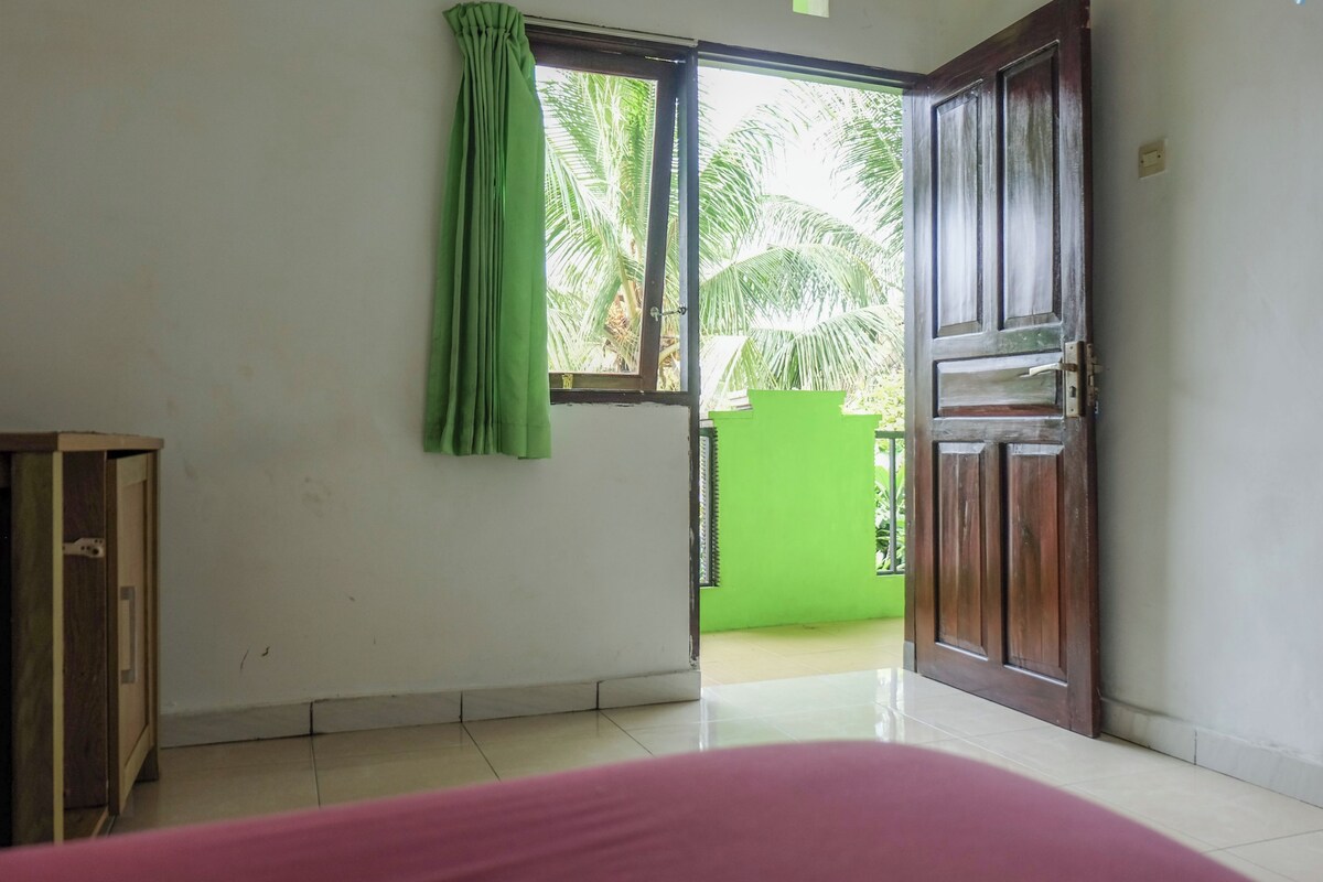 A room with a bed, a wooden door, a green curtain, and a view of palm trees outside.