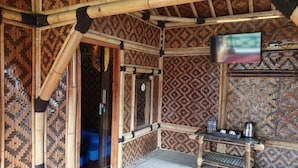Una habitación con pared de bambú con diseño, una cama, un televisor montado en la pared y una mesita con un hervidor y botellas.