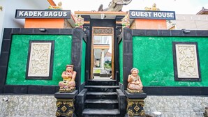 A building entrance with a sign reading "KADEK BAGUS GUEST HOUSE," flanked by two statues and a large dinosaur sculpture. - Bagus Kadek Guesthouse (Jimbaran)