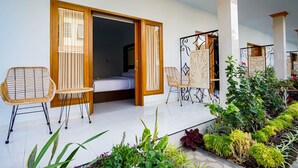 A room with a bed, two wicker chairs, a small table, and a view of a garden with various plants. - Bagus Kadek Guesthouse (Jimbaran)