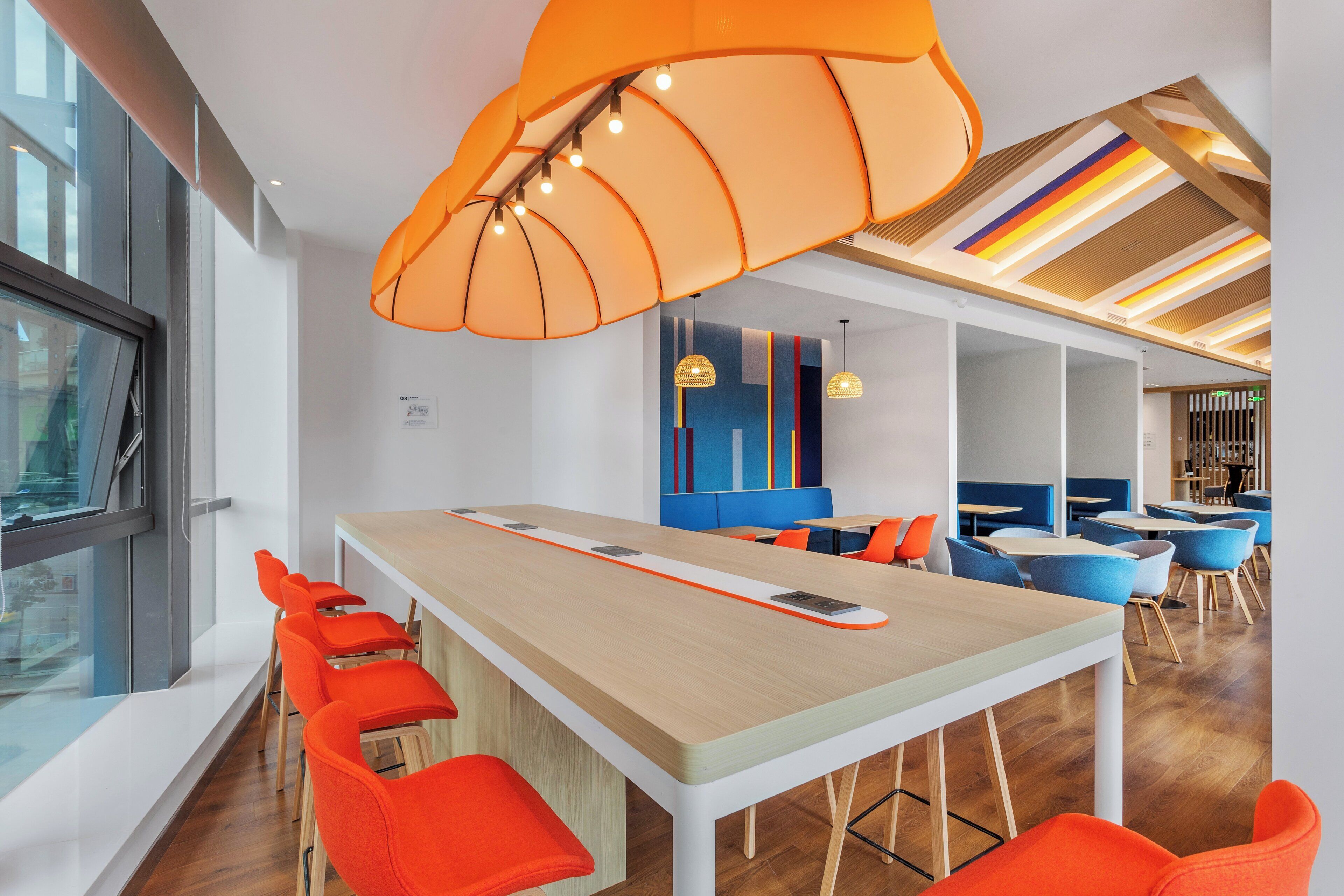 Un interior moderno con una larga mesa común, sillas color naranja brillante y un diseño de techo colorido.