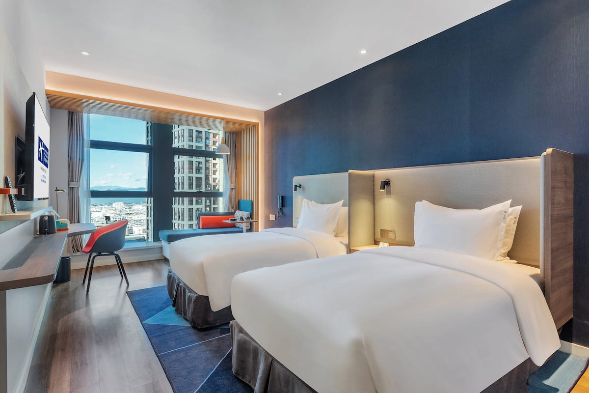 A hotel room with two beds, a desk, a chair, and a large window offering a city view.