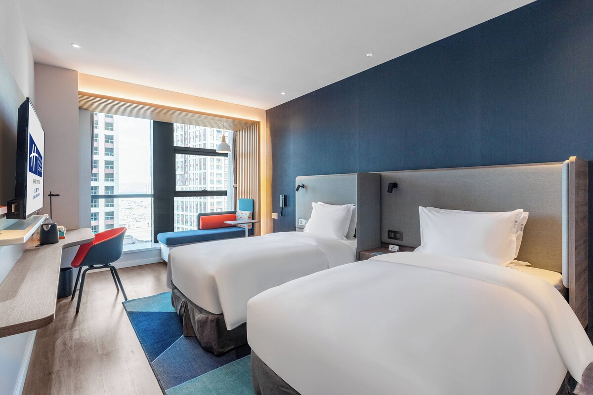 A hotel room with two beds, a desk, a chair, and a large window.