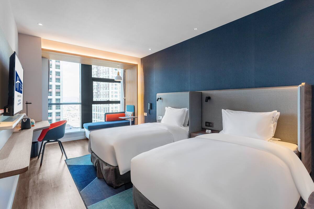 A hotel room with two beds, a desk, a chair, and a large window with a city view.