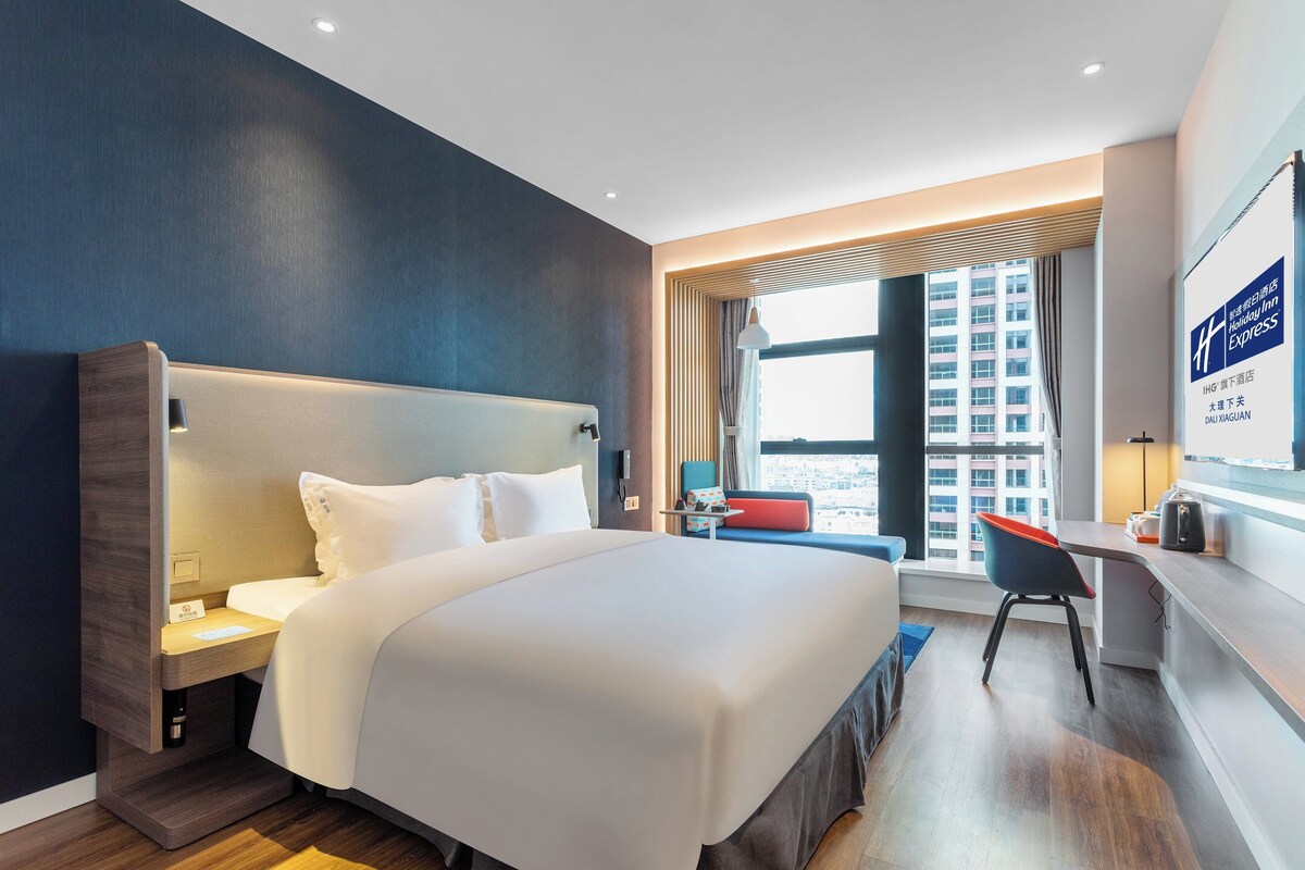 A modern hotel room with a large bed, a desk, and a view of the city.