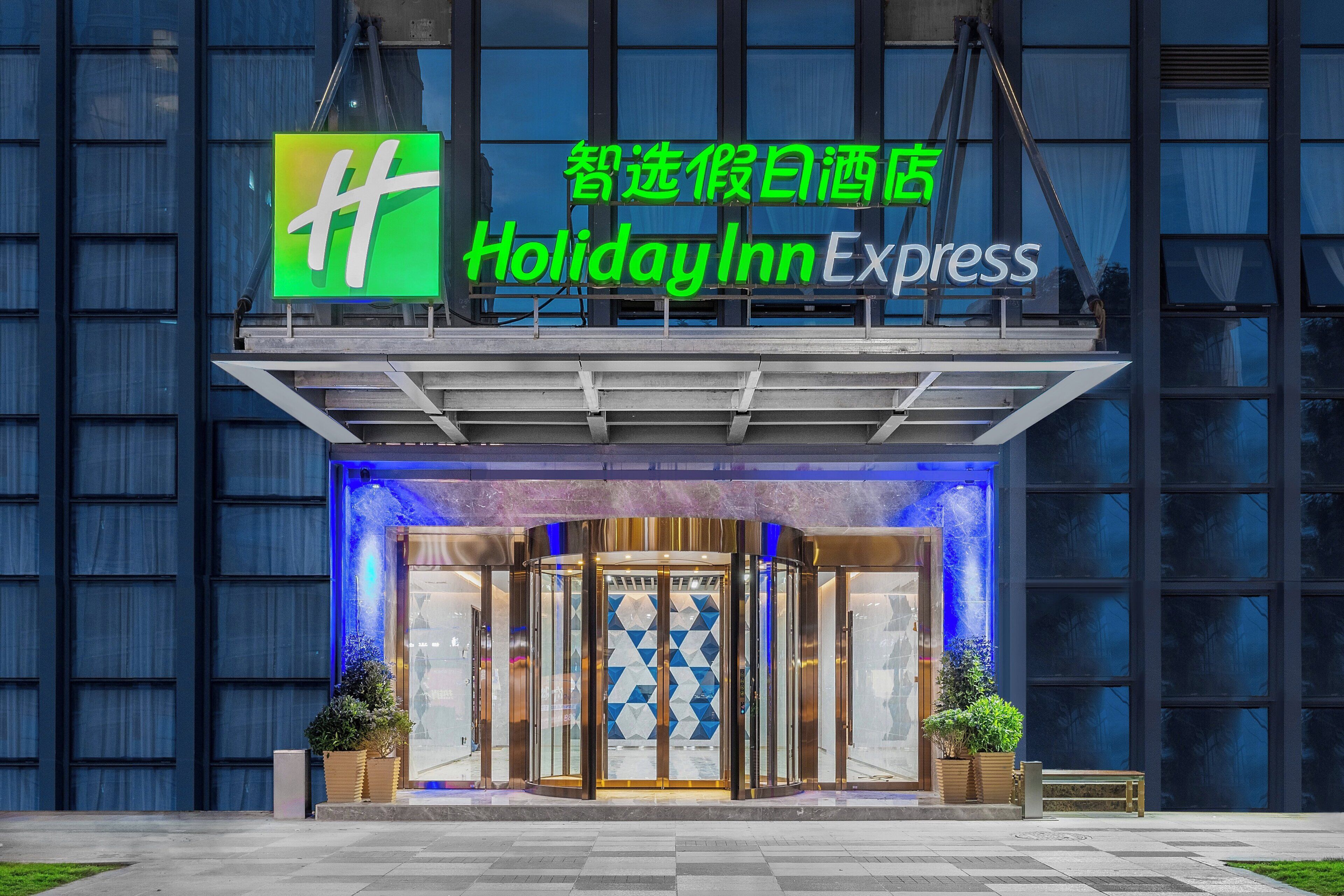 The exterior of a Holiday Inn Express hotel with a prominent green logo, a glass entrance, and a blue-lit facade.