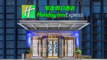 The exterior of a Holiday Inn Express hotel with a prominent green logo, a glass entrance, and a blue-lit facade.