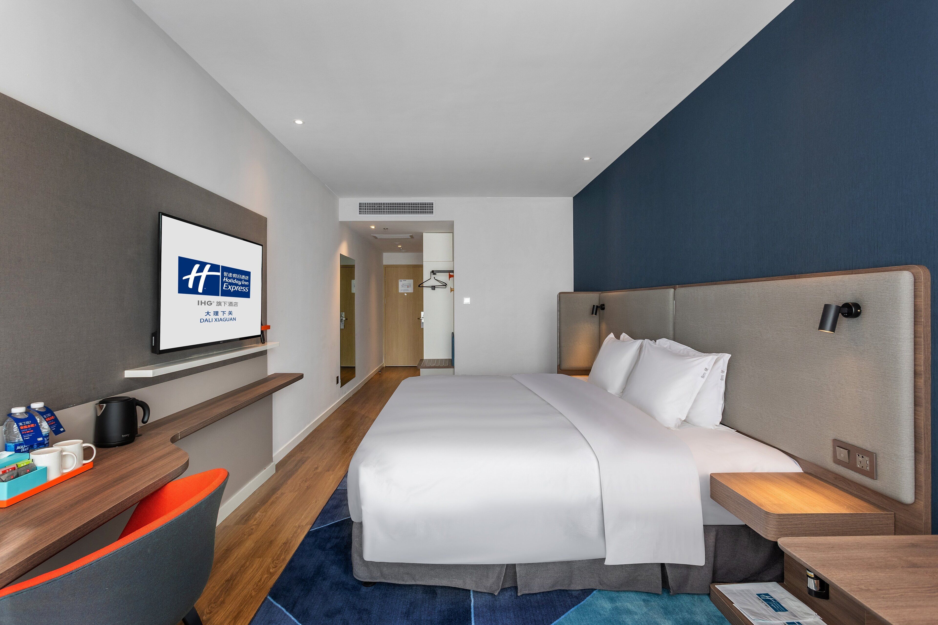 Quarto de hotel moderno com uma cama grande, uma escrivaninha com cadeira, televisão de tela plana e uma mesinha de cabeceira com abajur.