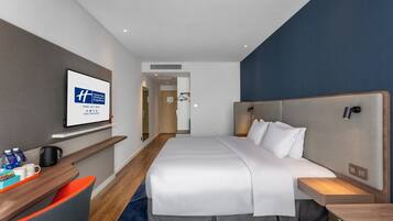 Quarto de hotel moderno com uma cama grande, uma escrivaninha com cadeira, televisão de tela plana e uma mesinha de cabeceira com abajur.