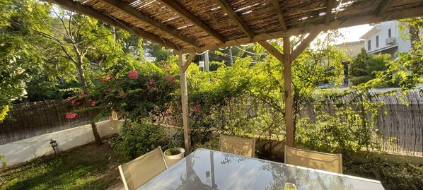 Une terrasse extérieure couverte, dotée d’une table et de chaises, et entourée d’une végétation luxuriante et de plantes en fleurs.