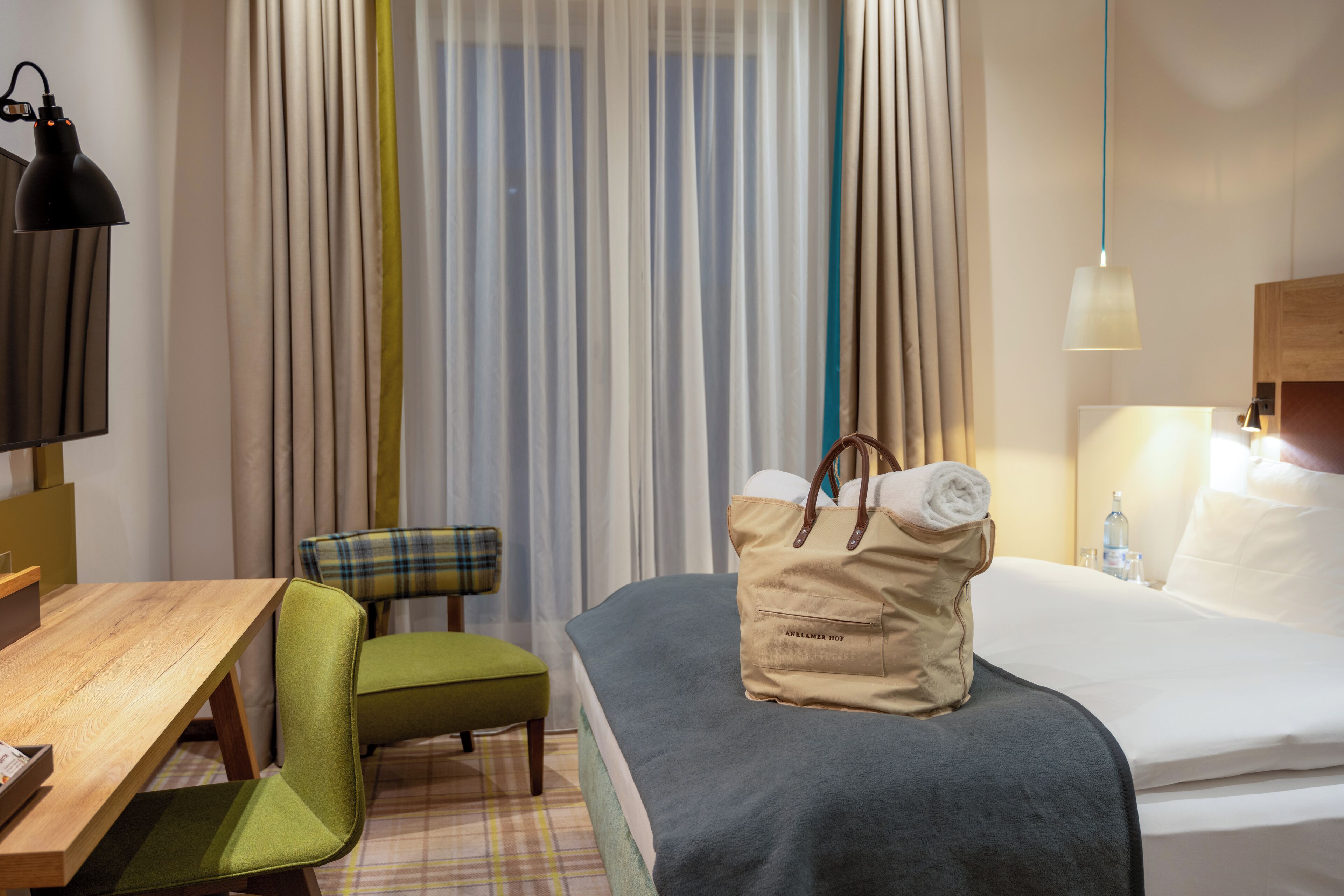 Habitación de hotel con cama, escritorio, silla, lámpara colgante y ventana con cortinas.
