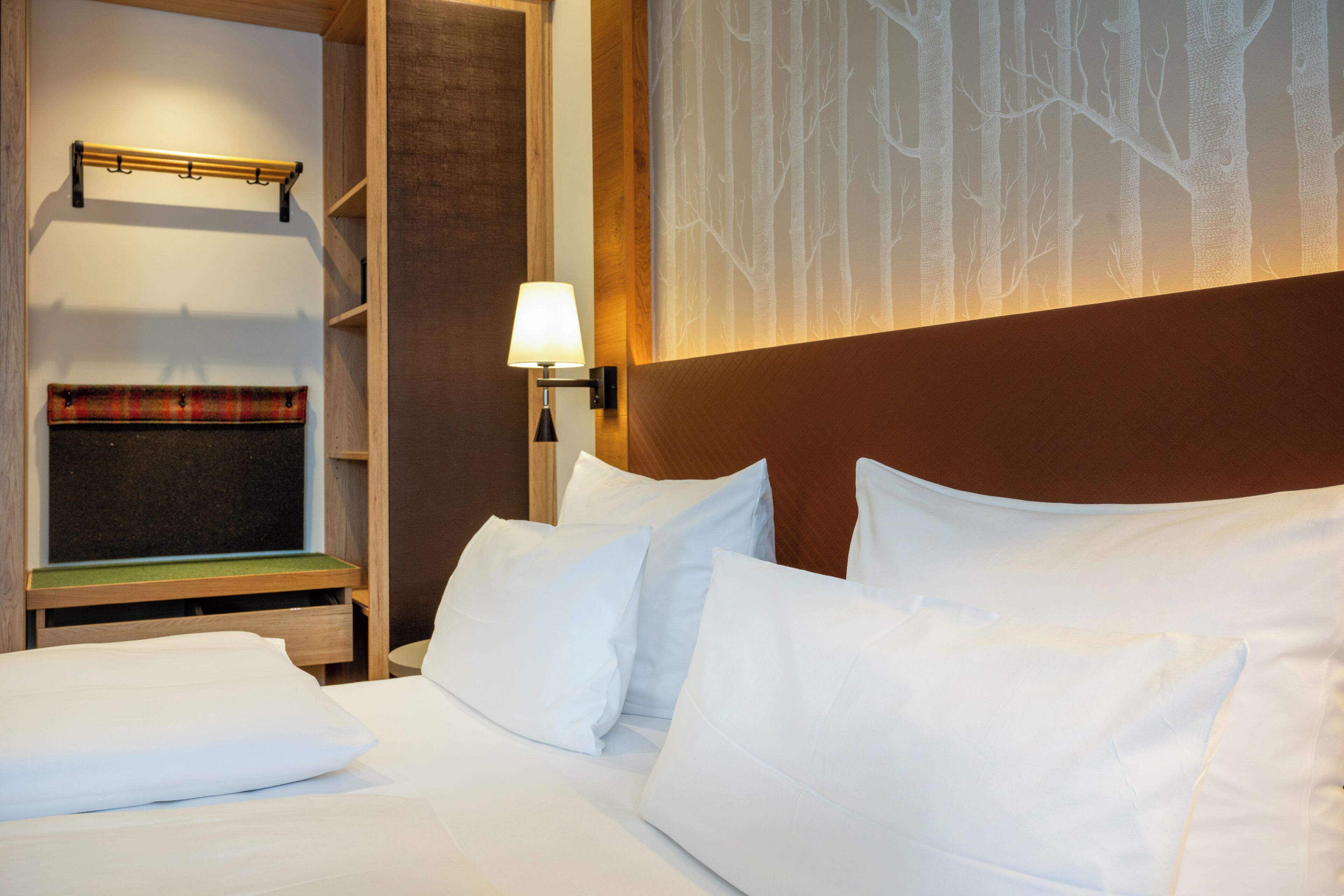 Une chambre d’hôtel avec un lit, des oreillers blancs, une table de chevet avec une lampe, un porte-manteau et un mur orné d’un motif représentant un arbre.