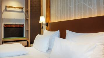 Une chambre d’hôtel avec un lit, des oreillers blancs, une table de chevet avec une lampe, un porte-manteau et un mur orné d’un motif représentant un arbre.