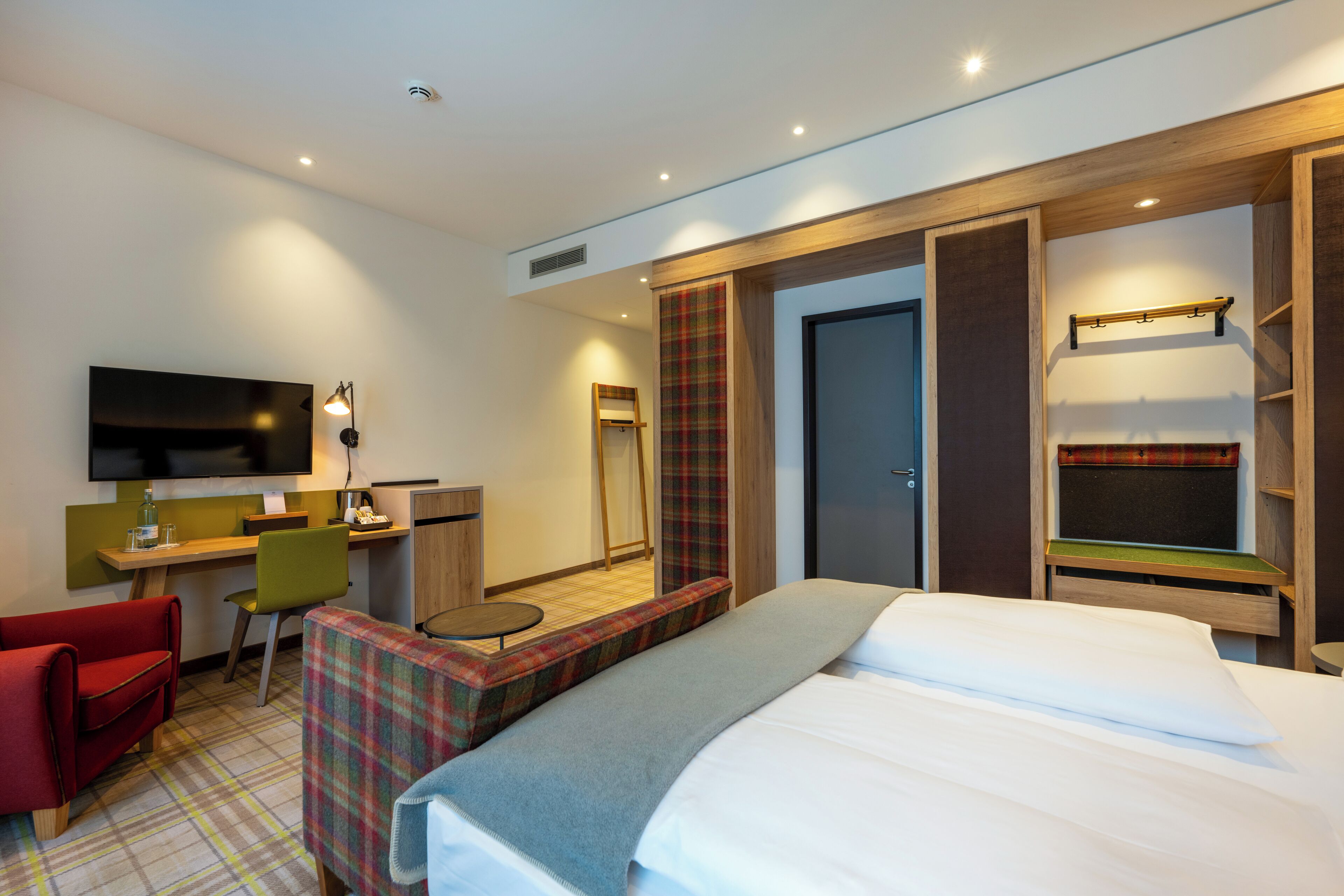 A modern hotel room with a bed, a desk, a chair, a TV, and a wardrobe.