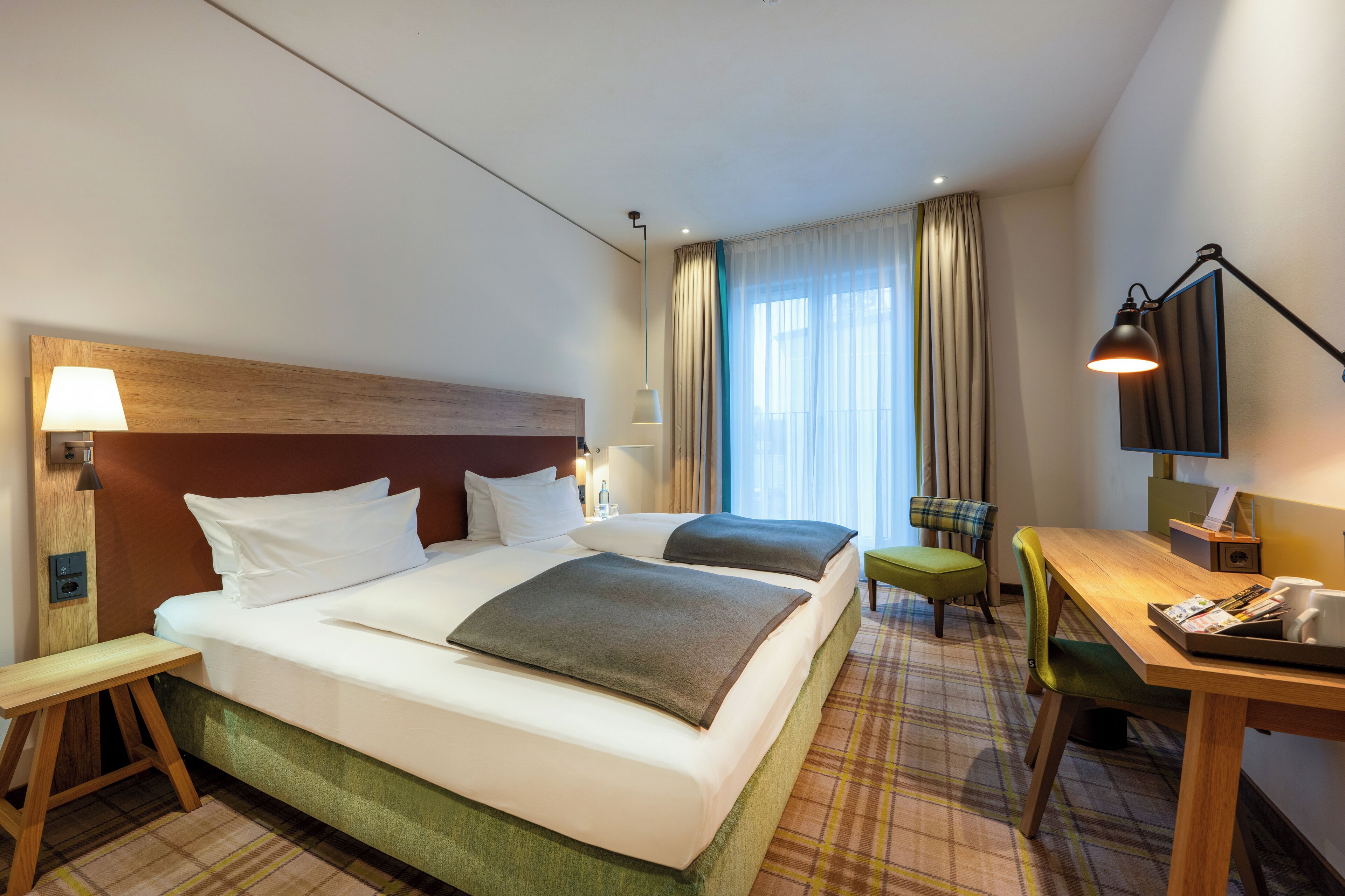 Une chambre d’hôtel comprenant un lit, deux tables de chevet, un bureau, une chaise, une lampe et une fenêtre avec des rideaux.