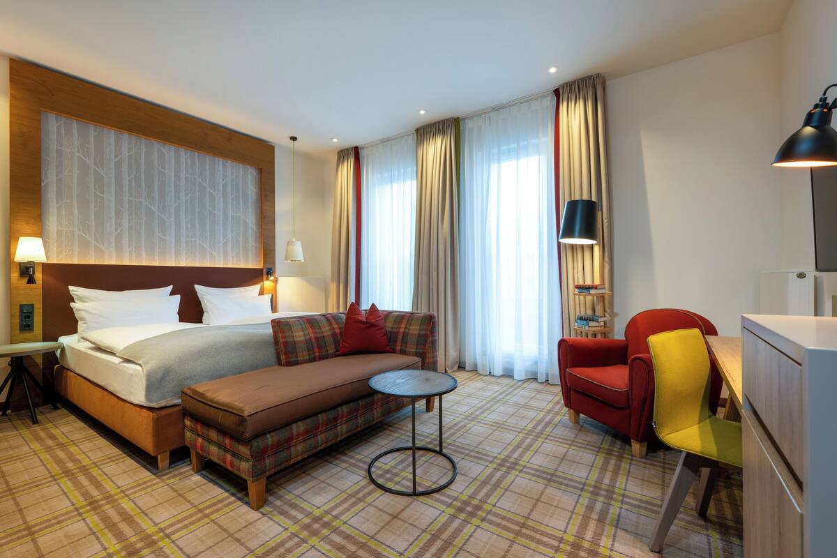 Habitación de hotel con cama, sofá, sillón, pequeña mesa redonda y escritorio con lámpara.