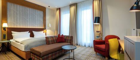 Habitación de hotel con cama, sofá, sillón, pequeña mesa redonda y escritorio con lámpara.