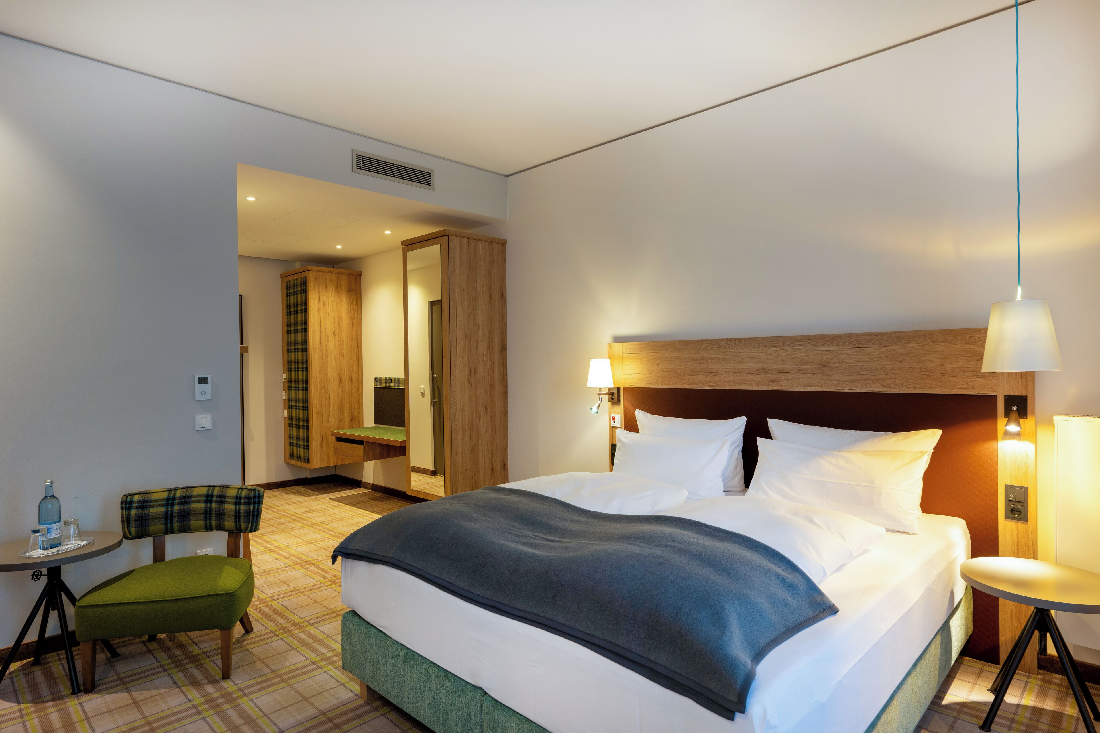 A hotel room with a large bed, a small table with a chair, a mirror, and a wardrobe.