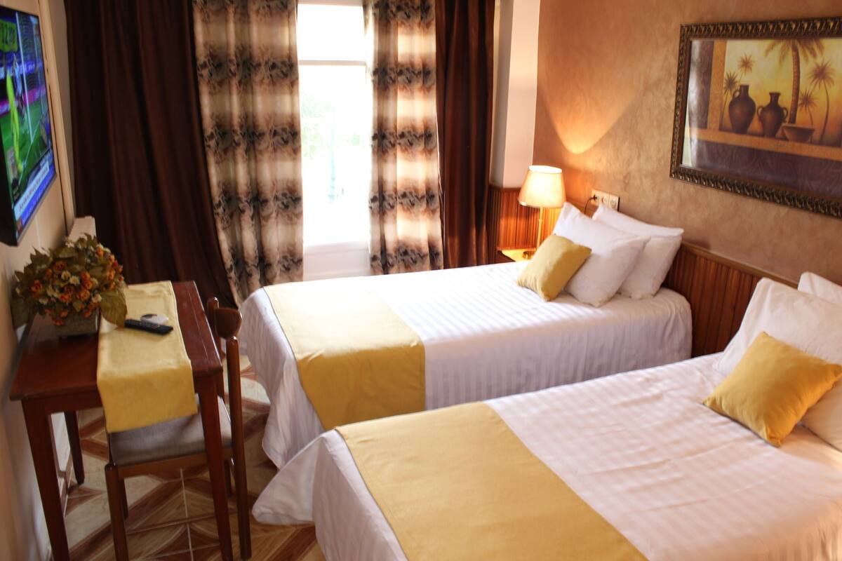 A hotel room with two beds, a desk with a chair, a TV, and a painting on the wall.