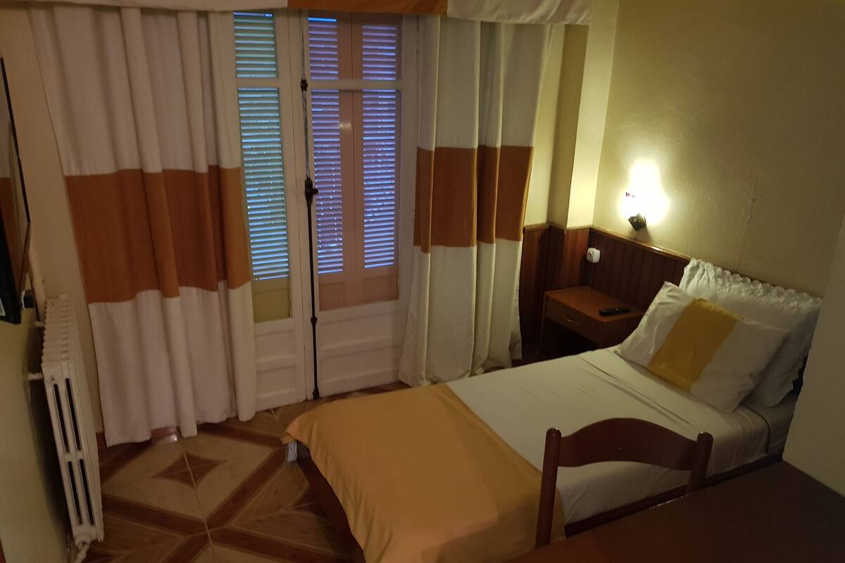 A hotel room with a bed, a nightstand, a table, and a radiator.