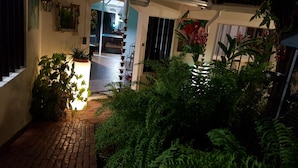 A narrow brick pathway leading to a pool area with a glass door, surrounded by lush greenery and a hanging light fixture.