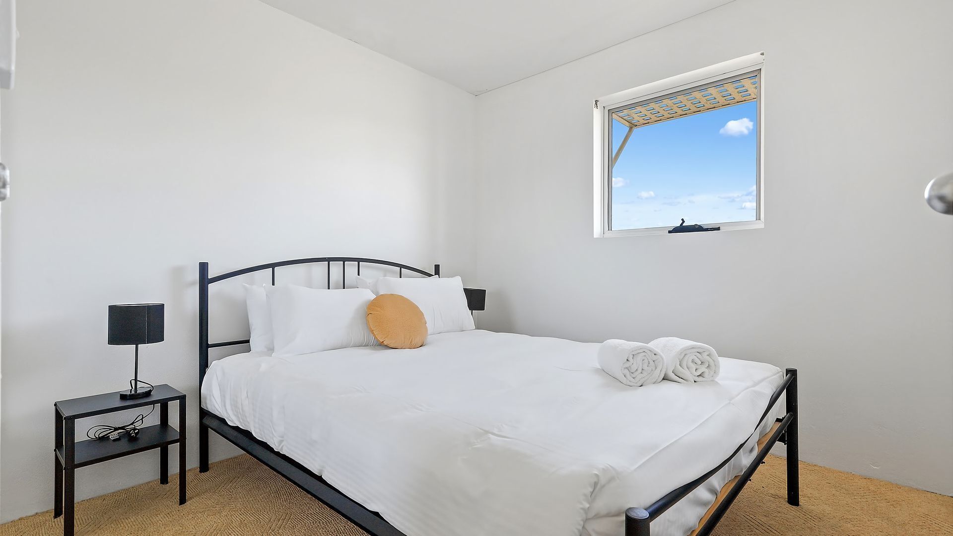 A bedroom with a bed, a nightstand, a lamp, and a window with a view of the sky.