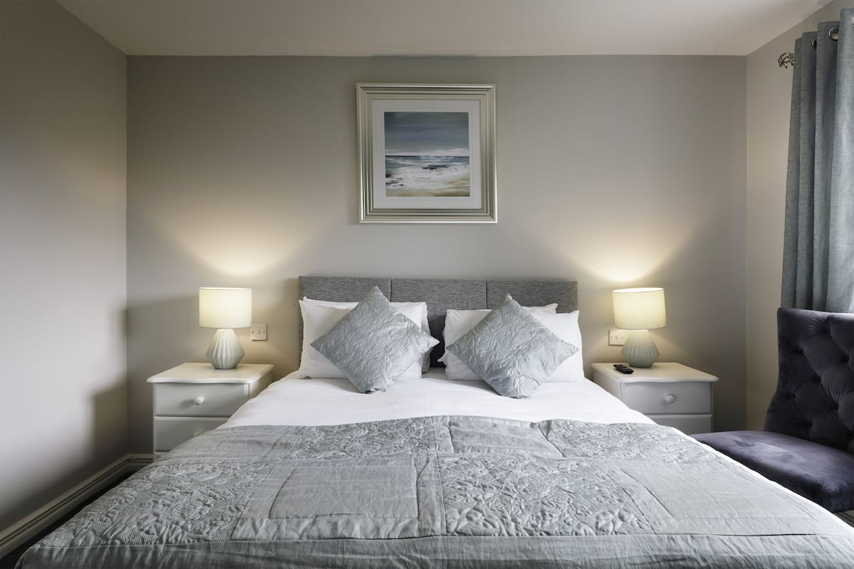 A neatly made bed with a grey quilt, two bedside tables with lamps, a grey upholstered chair, and a framed picture on the wall.